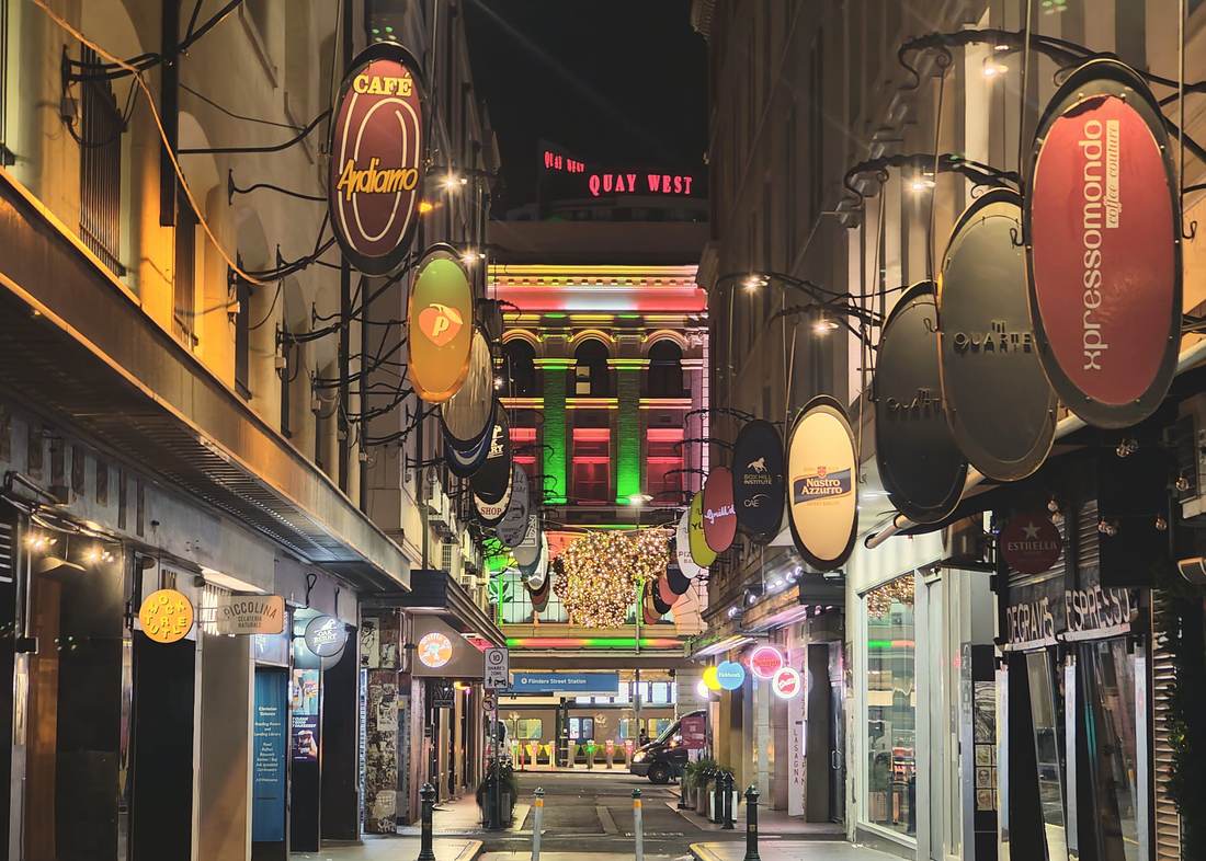 A narrow city laneway at night, hung with round illuminated café signs on both sides and lit shopfronts below.