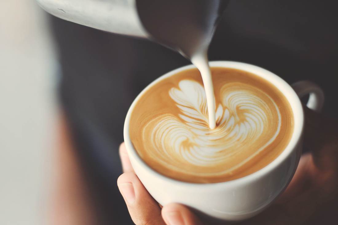 A barista pouring steamed milk into a coffee, a leaf of latte art forming in the cup.