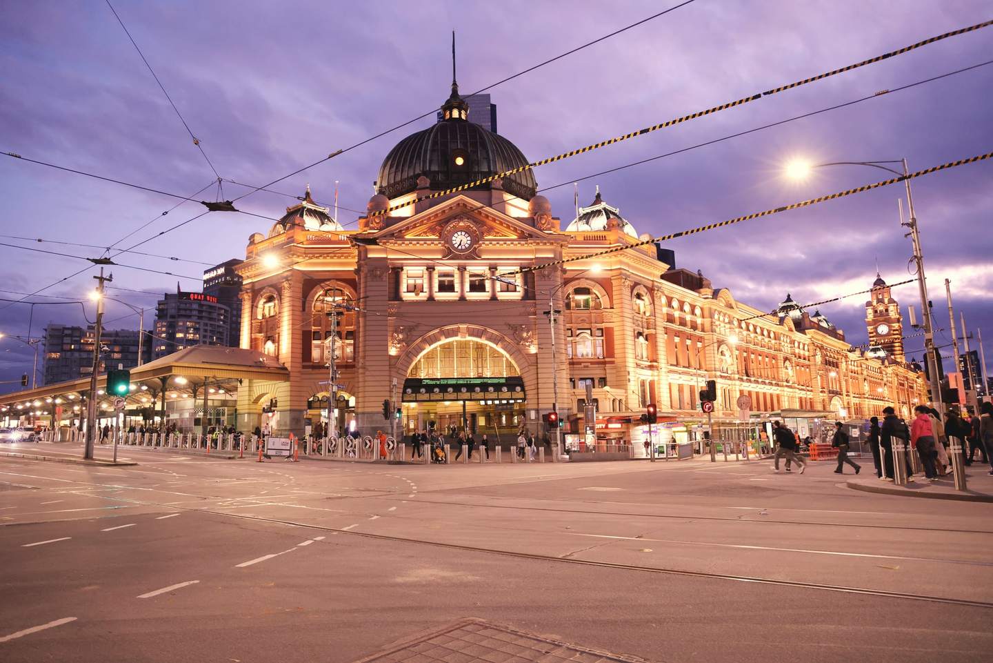 Flinders Street Station lit at dusk, its dome and clocks above the entrance, tram wires crossing the intersection in front.