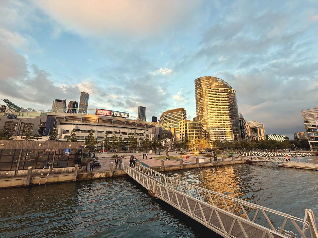 A stadium with a closed roof beside the water at Docklands at sunset, city towers behind it and boats moored in front.