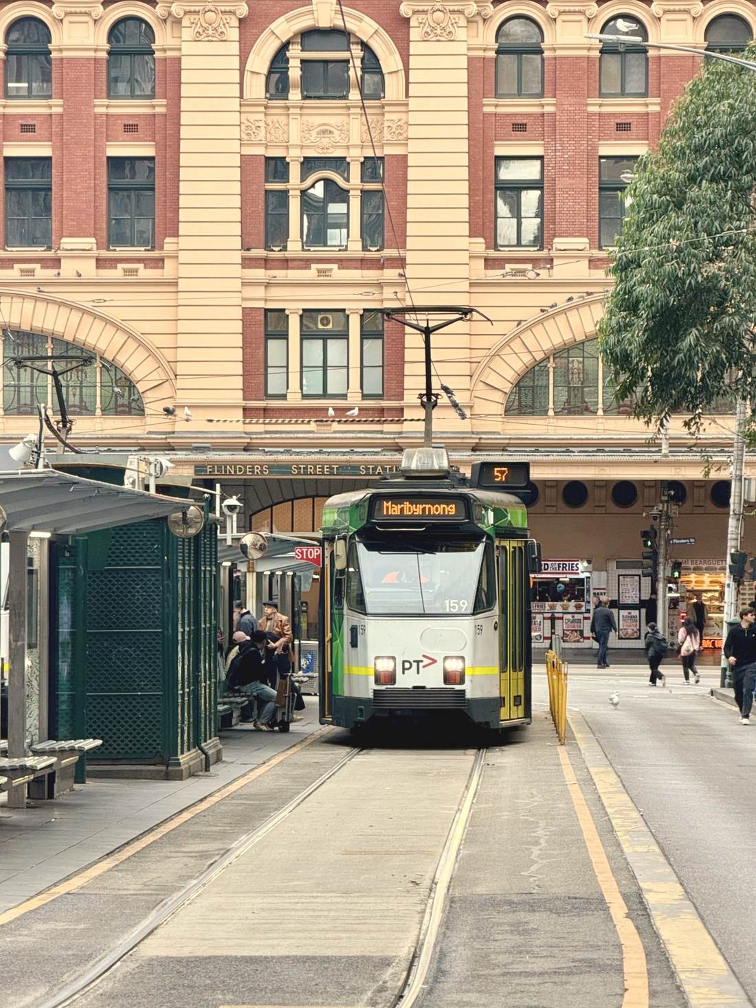 A green and yellow tram stopped at a platform in front of Flinders Street Station, with passengers waiting under the shelter.