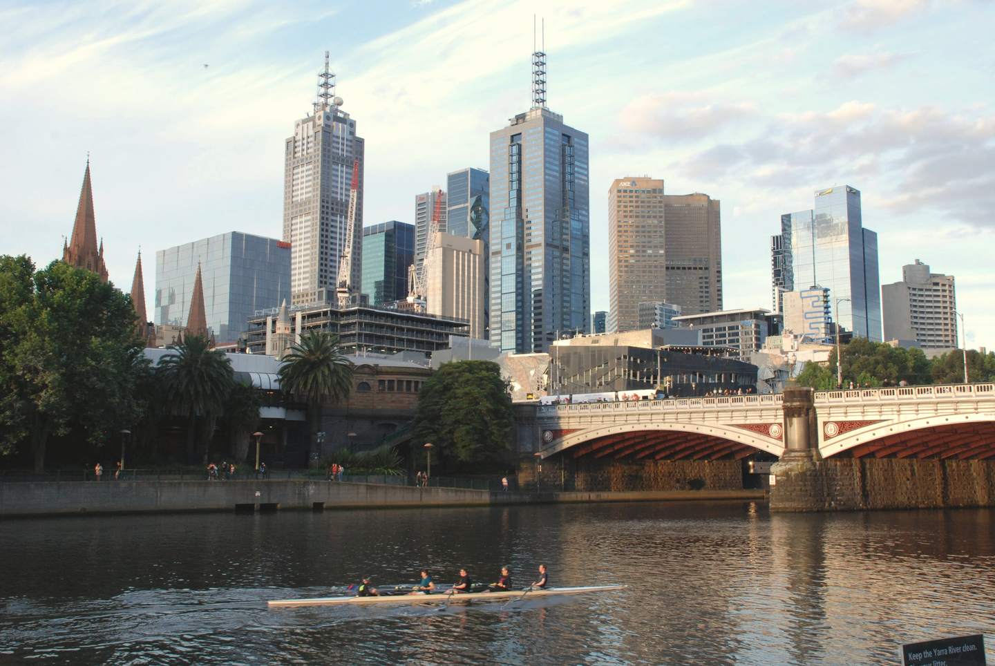 Princes Bridge in late-afternoon sun, a rowing crew on the Yarra in the foreground and the city towers behind.