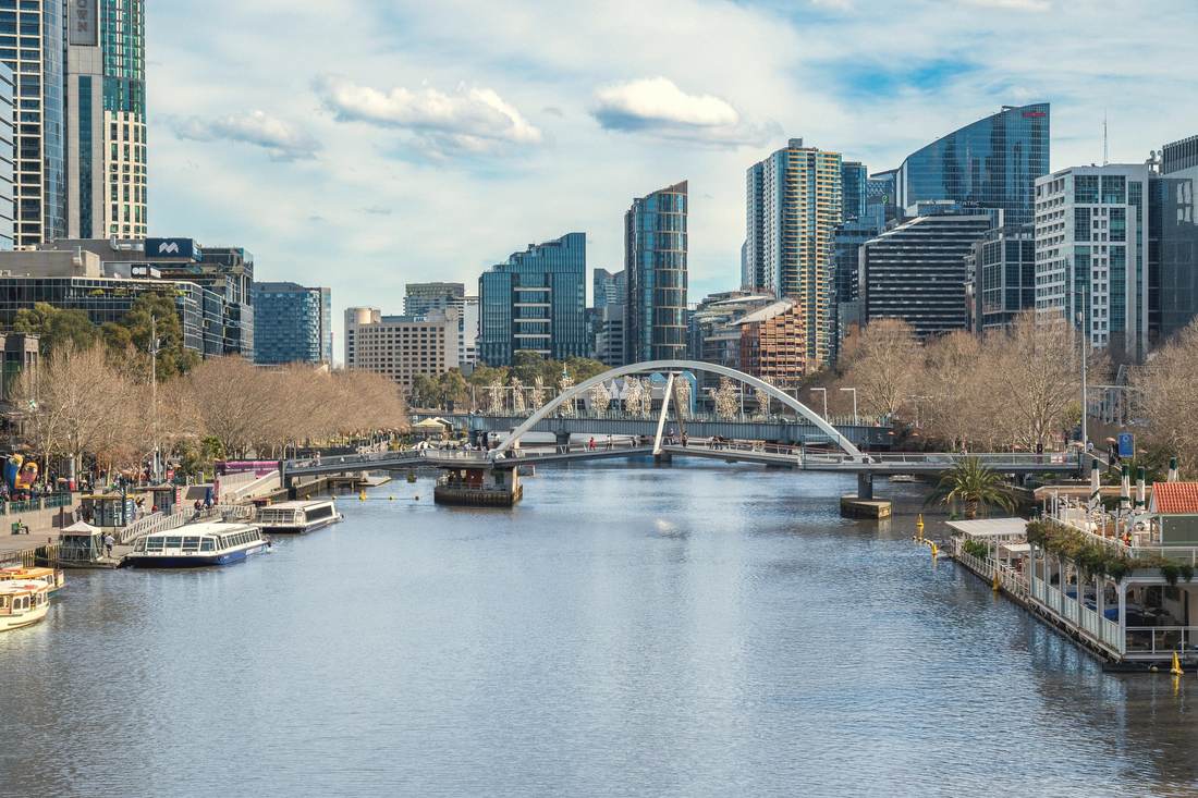 The Southbank promenade on a clear day, walkers along the river, cruise boats moored at the edge and a white footbridge crossing the Yarra.