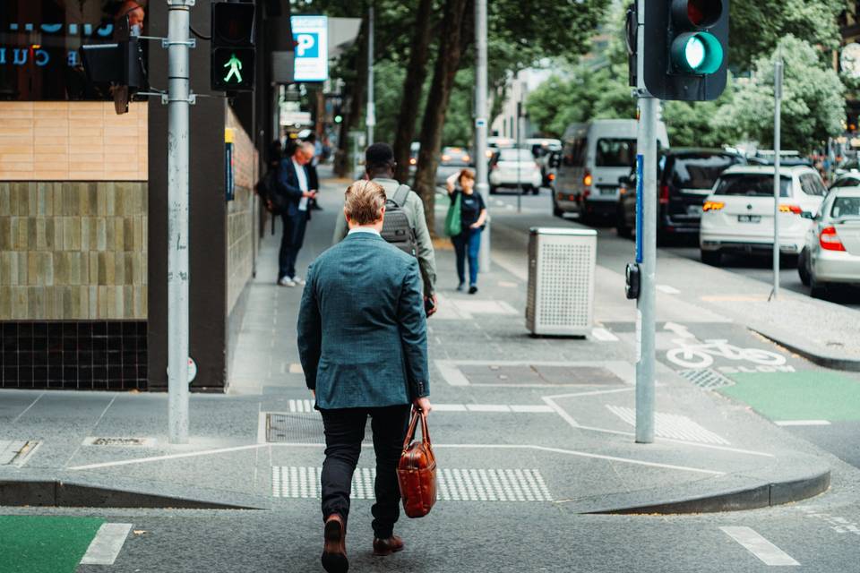 A man in a blue suit crosses a tree-lined street in the Melbourne CBD on a bright day, bag in hand.