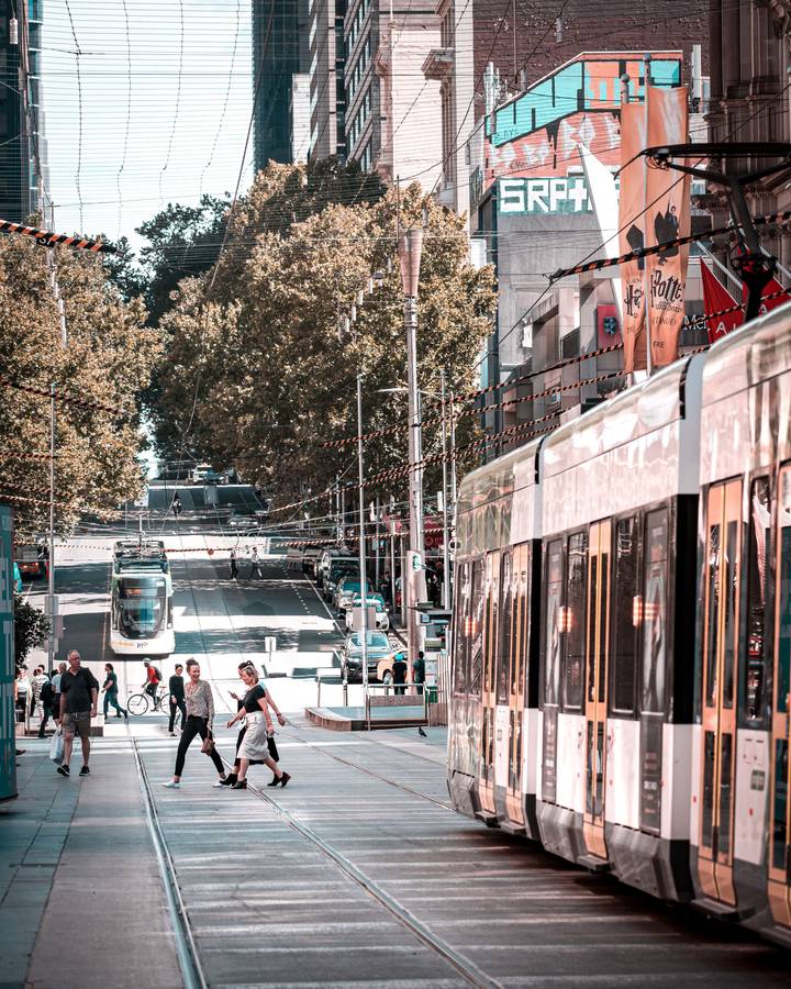 Pedestrians crossing a Melbourne city street beside a tram on a bright day.