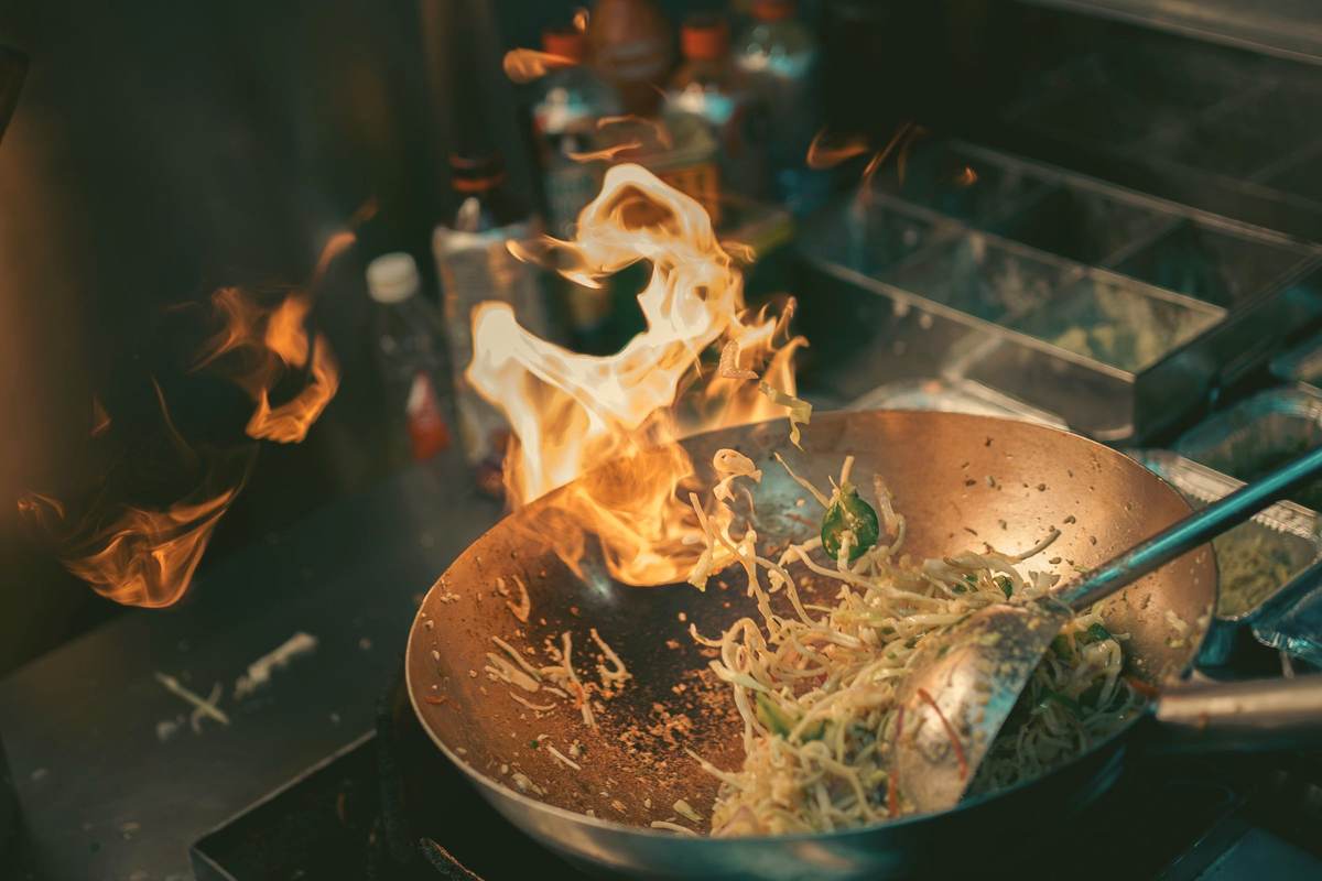 Noodles tossed in a wok over open flame in a restaurant kitchen.