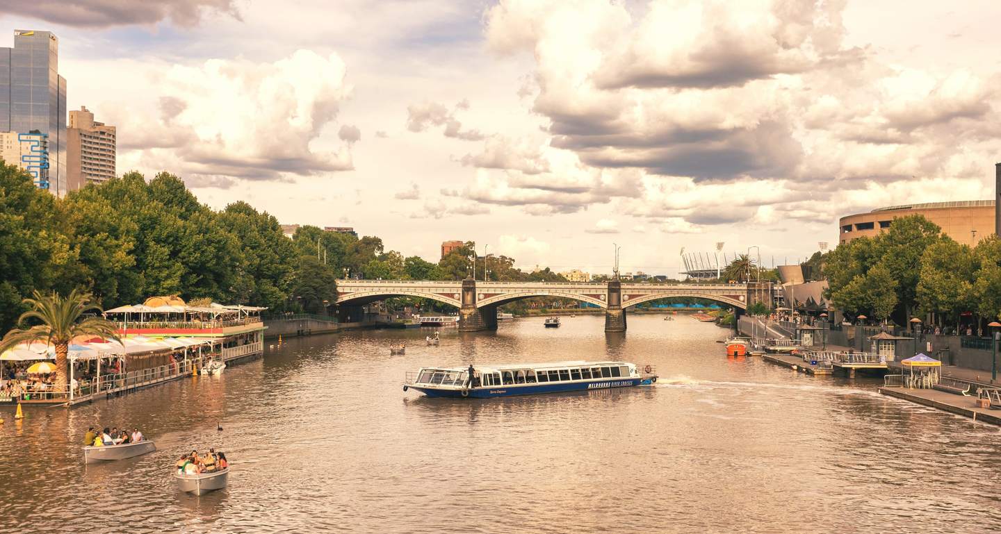 The Yarra River on a bright afternoon, rowing boats and a river-cruise boat on the water, umbrellas along the north bank and Princes Bridge ahead.